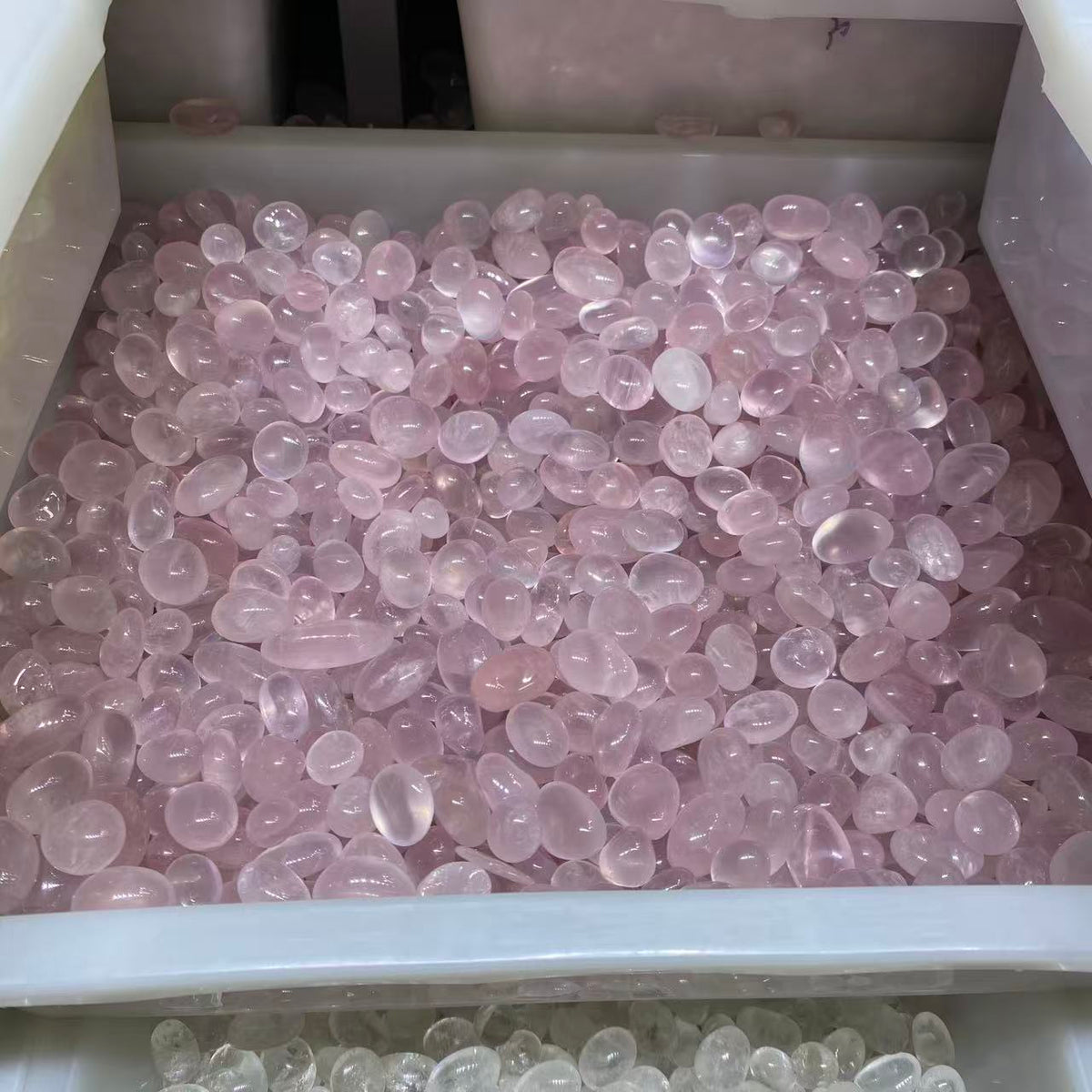 A large white plastic storage bin filled with many small, smooth, translucent pink stones or beads, photographed from above. A second drawer or bin at the bottom edge shows pale clear stones.