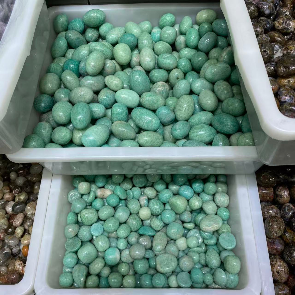 A bulk display of polished river stones, with two white plastic bins filled with light green mottled stones and bins of brown stones partially visible on the sides.
